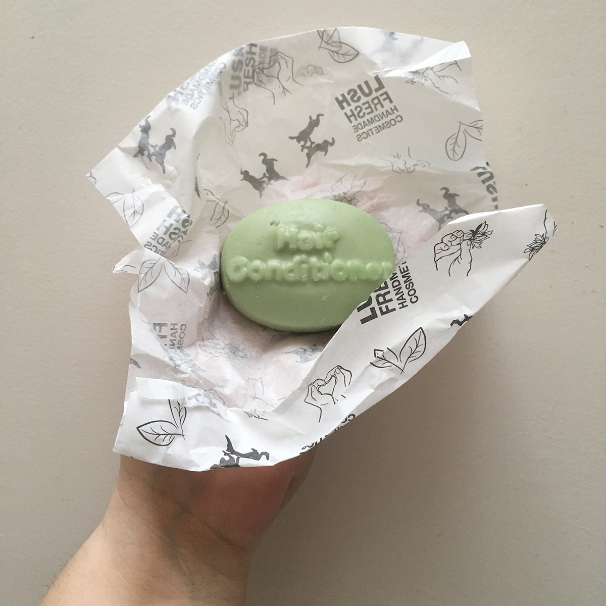 A hand holds up a bar of Lush Jungle Solid Conditioner wrapped in paper