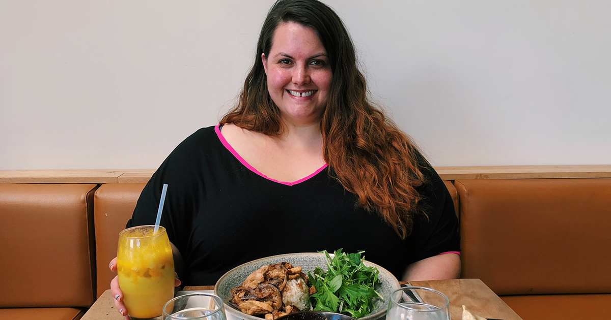 Meagan sits at Wagamama restaurant wearing a black dress with hot pink piping around the collar. In front of her is a table with various dishes of food