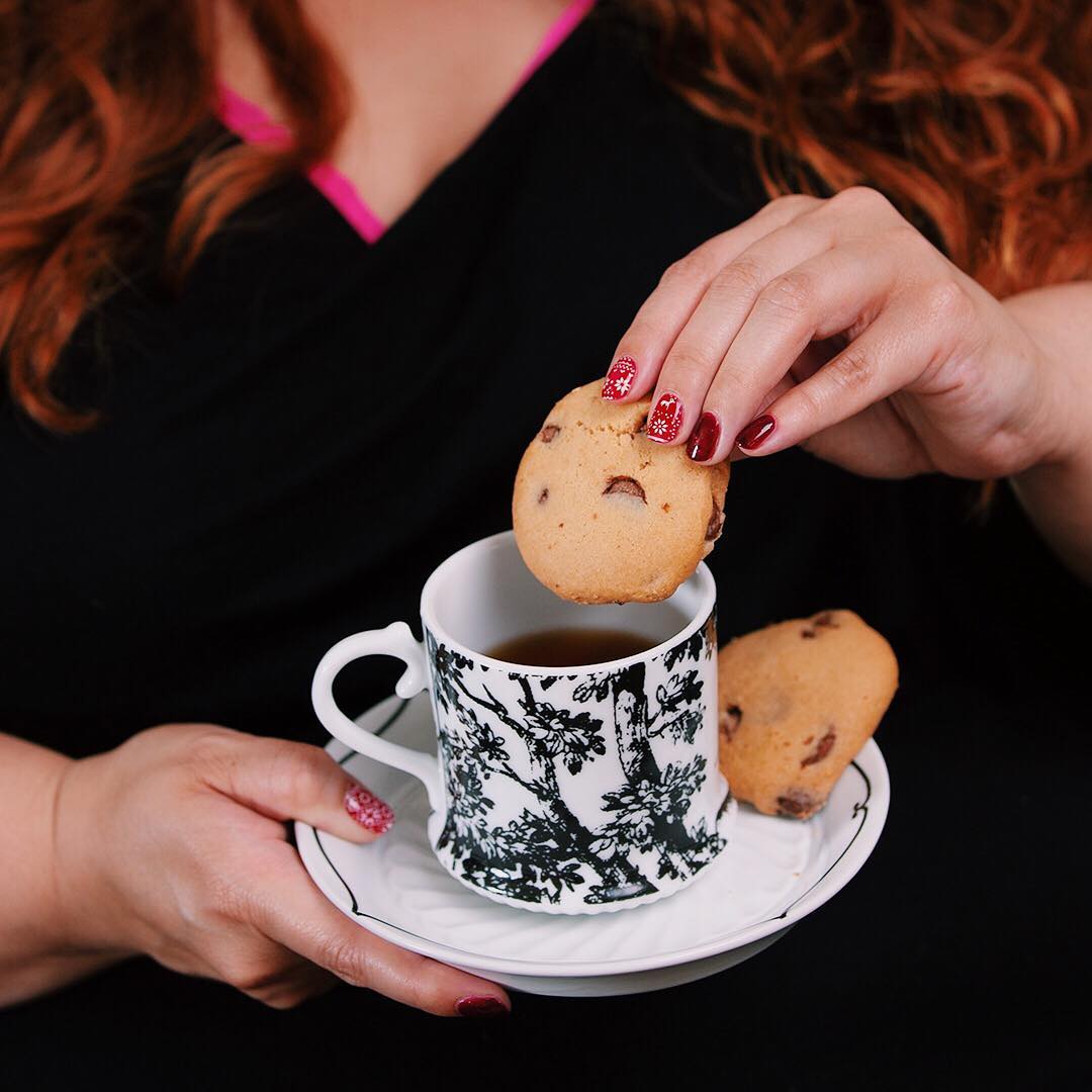 A close up of Meagan holding a cup and saucer in her right hand. With her left hand she is about to dunk a chocolate chip cookie in her tea, and another cookie rests on the saucer