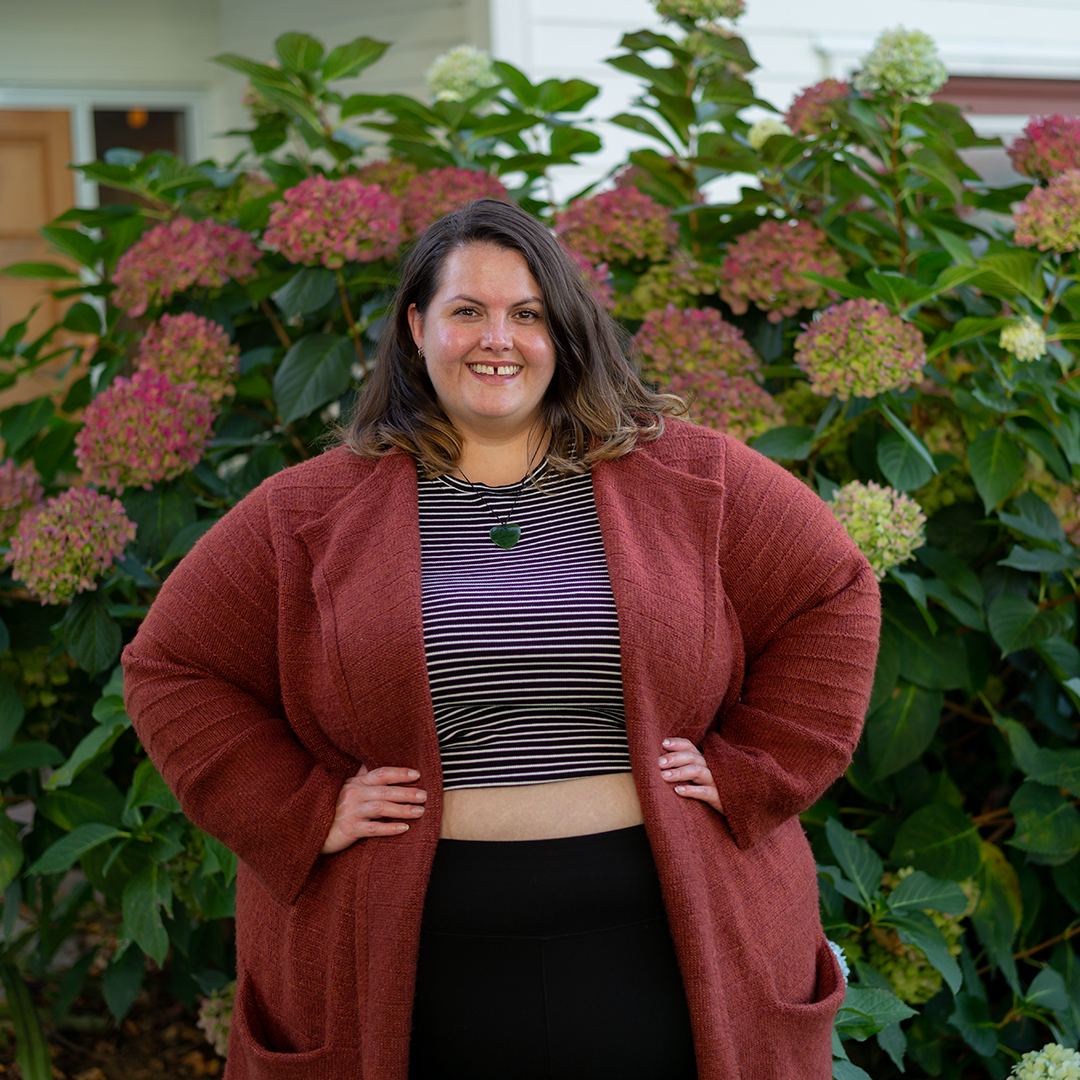 New Zealand plus size blogger Meagan Kerr stands in front of a hydrangrea bush. The photo shows her from her hips upward, and she is standing with her hands on her waist. Meagan wears a midriff baring City Chic Cheeky Stripe Top in XXL, Snag Tights Leggings in H, Old Navy Rust Cardigan in 4X