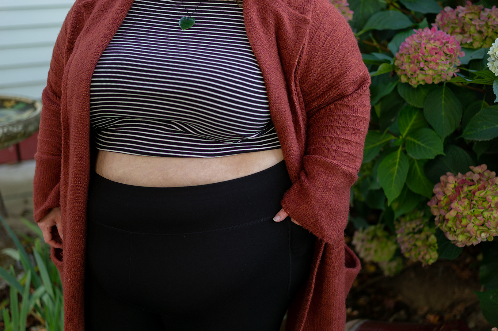 New Zealand plus size blogger Meagan Kerr stands in front of a hydrangrea bush. The photo is cropped to show from just below her chin to her hips, and she is standing with one hand in the pocket of her leggings. Meagan wears a midriff baring City Chic Cheeky Stripe Top in XXL, Snag Tights Leggings in H, Old Navy Rust Cardigan in 4X