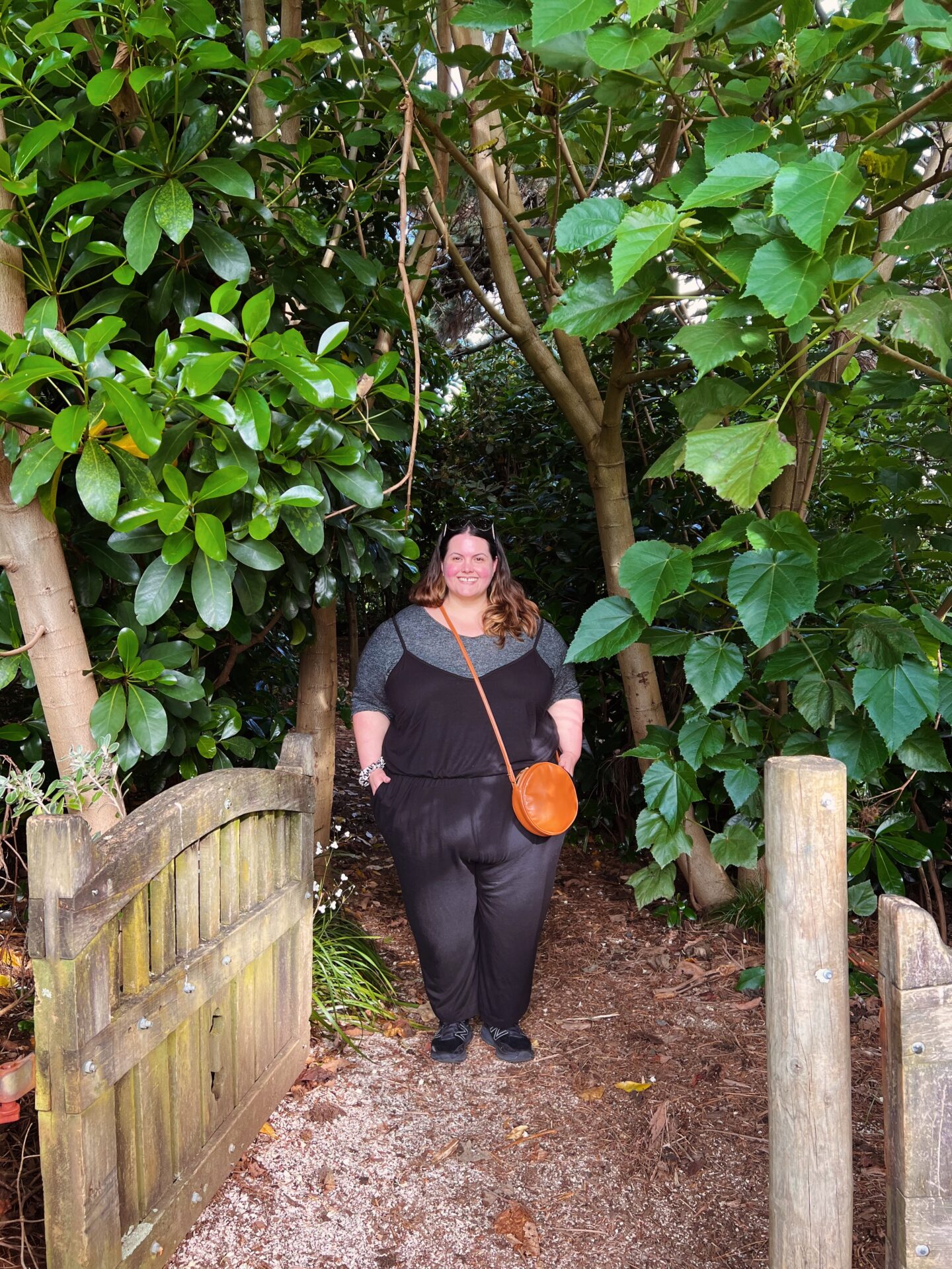 Meagan, a fat Māori woman, stands at the gated entrance to a wooded area. She is wearing a black jumpsuit with spaghetti straps over a grey marle tee, black sneakers and a tan cross body bag.