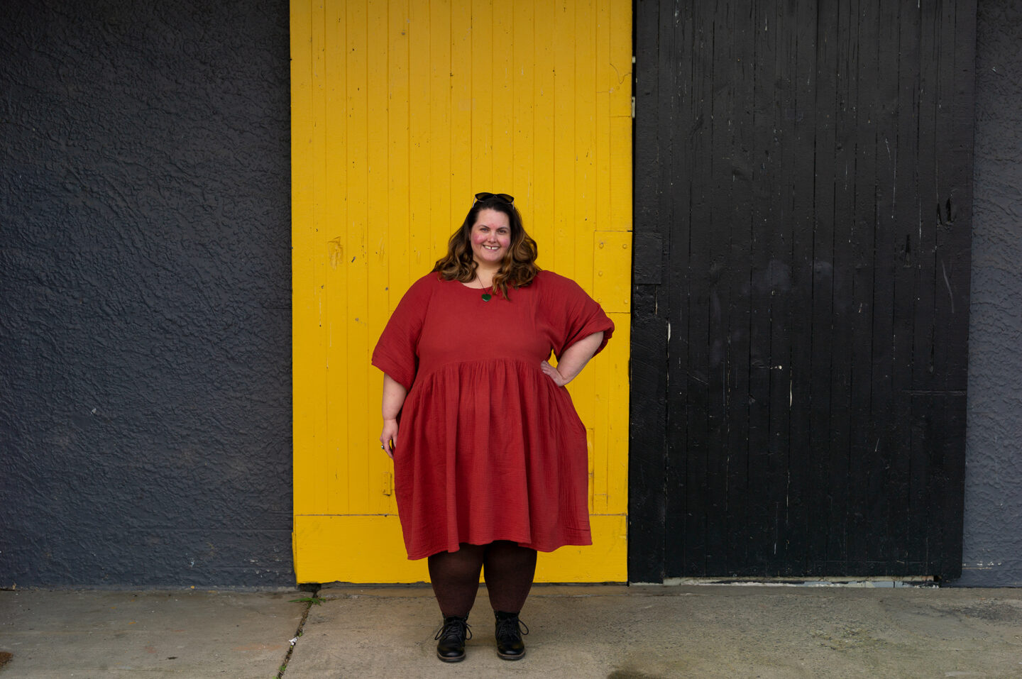 Meagan, a fat Māori woman, stands in front of a yellow door in a black wall. She is wearing a brick red dress with brown marl stockings and black lace up boots.