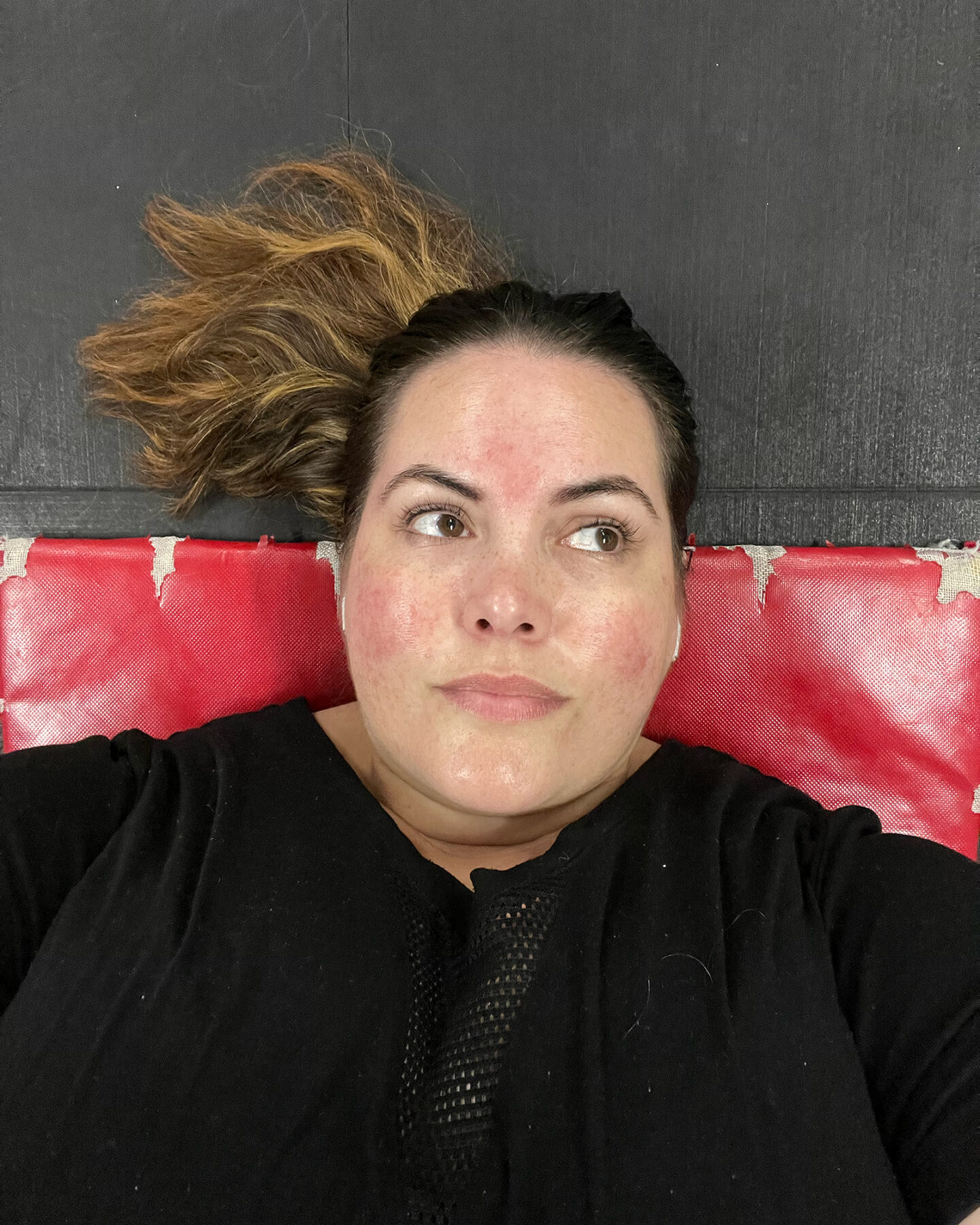 A selfie of fat Māori woman with lipoedema lying on the floor of the gym on a worn red mat. She is looking up, away from the camera, and her hair is fanned out in a ponytail on the floor.