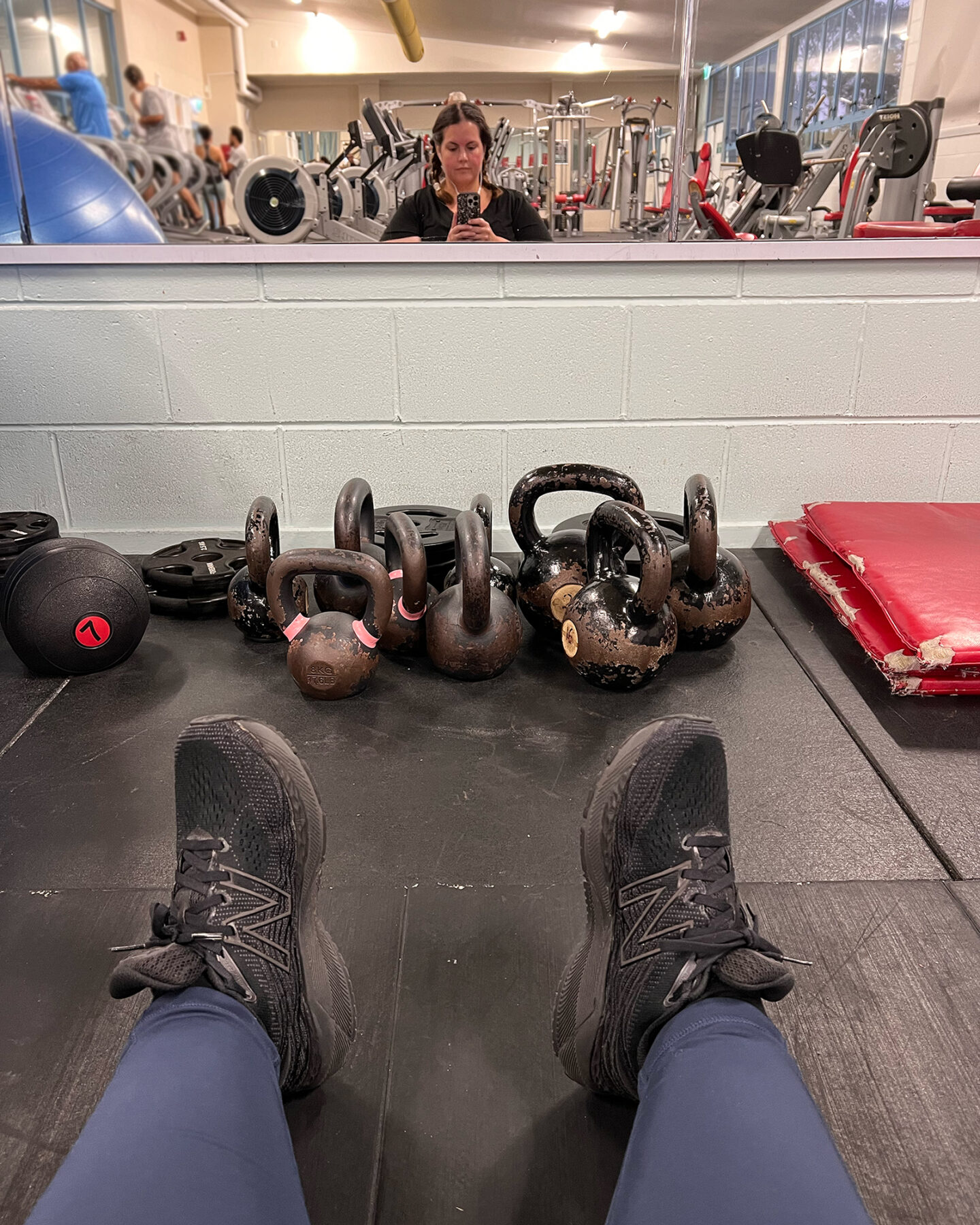 A fat Māori woman with lipoedema is sitting on the floor at the gym. She is taking a selfie in a mirror. Her feet are sticking out in front of her, and there are kettlebells piled on the floor between her and the mirror.