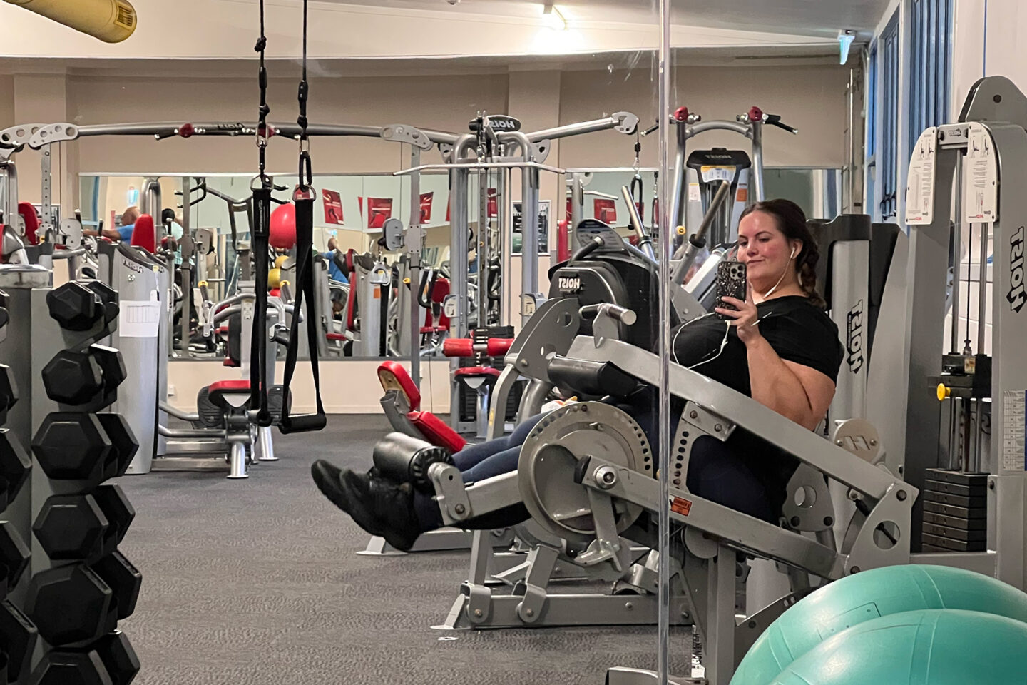 A fat Māori woman with lipoedema is sitting on a leg curl and extension machine in an empty gym. She is taking a selfie in a mirror.