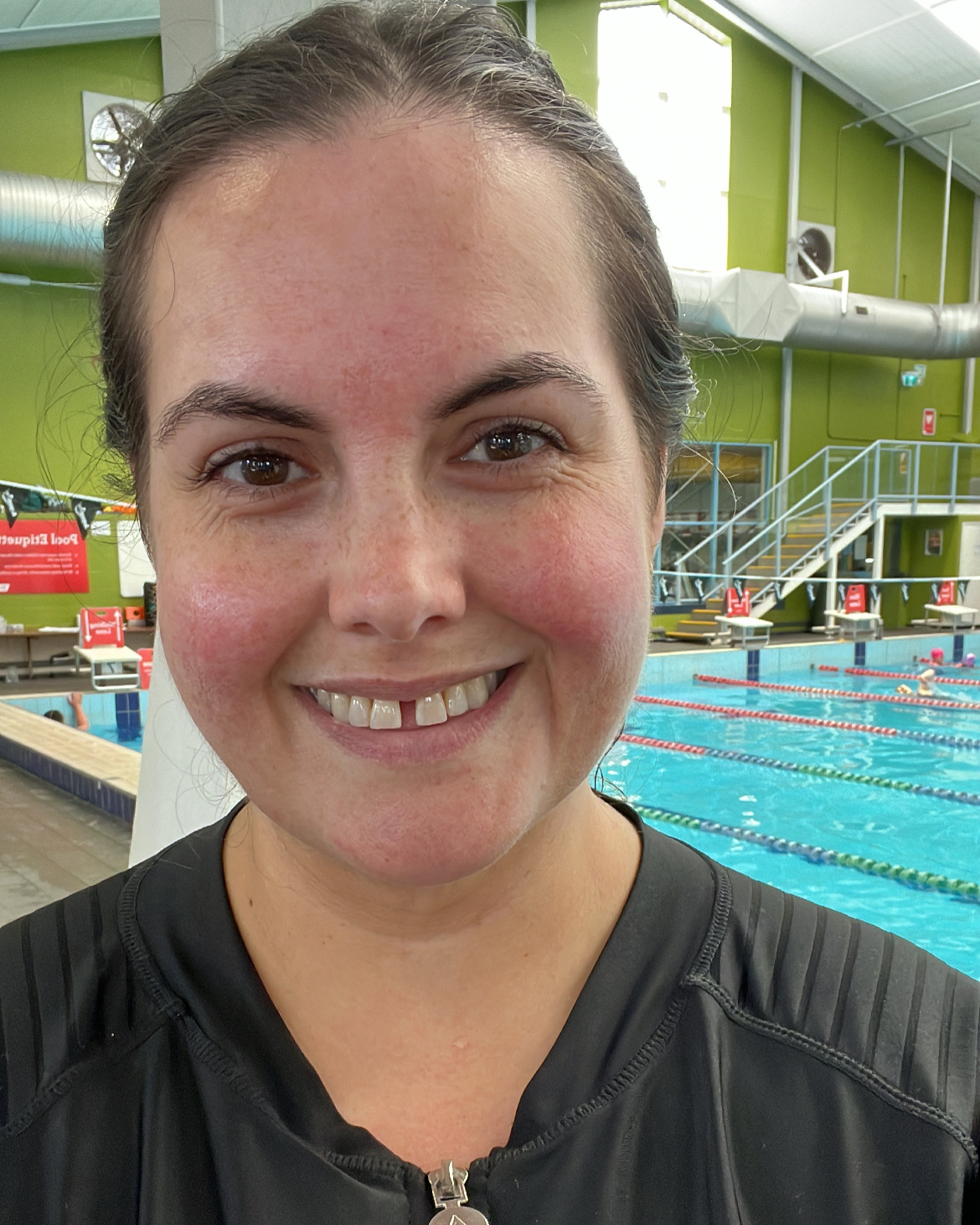 A selfie of a Māori woman at an indoor swimming pool. She is wearing a rash shirt and is smiling at the camera.