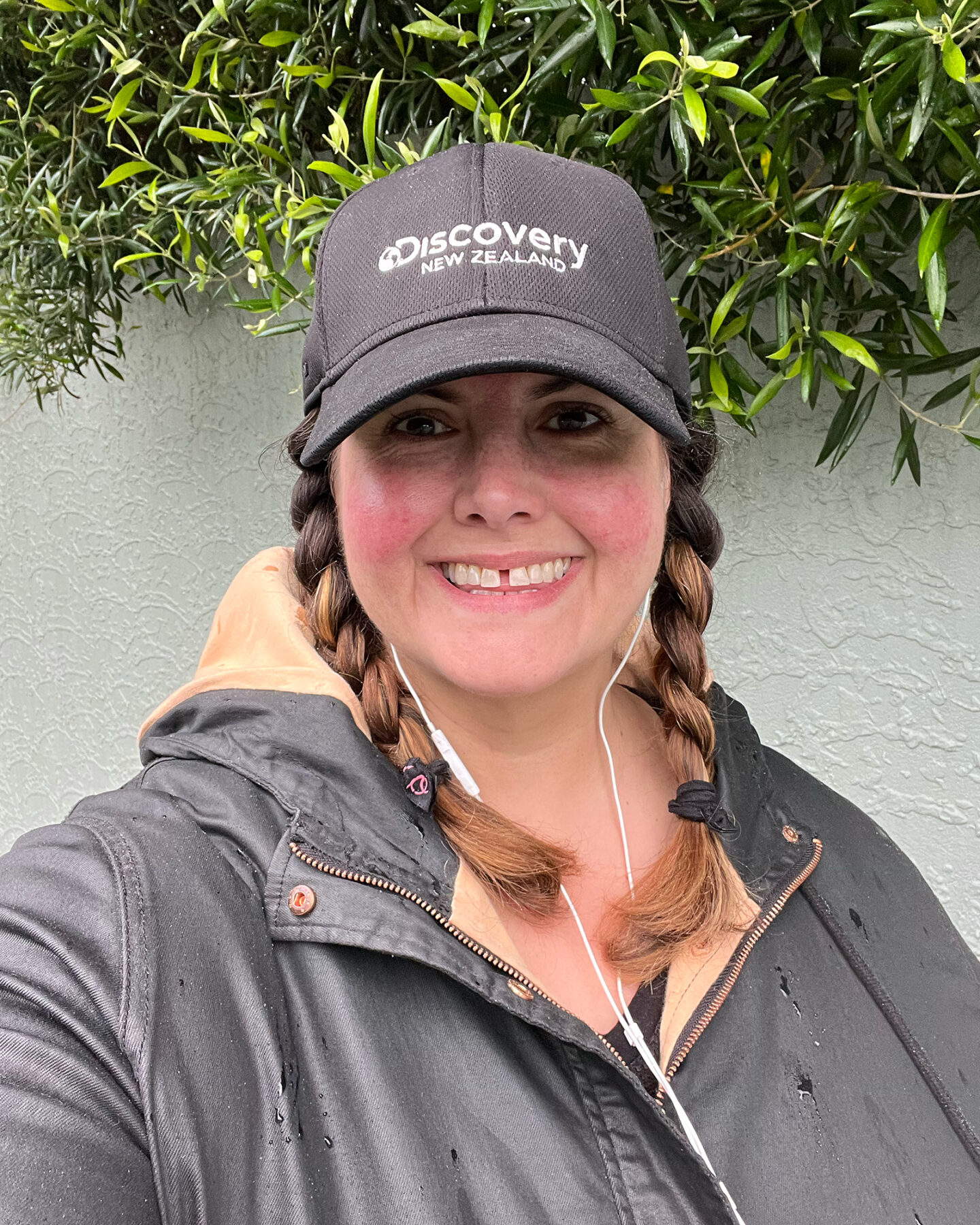 A selfie of a Māori woman walking outside in front of a light green wall with olive trees growing over the top. She is wearing a black rain jacket with a black cap that says Discovery New Zealand and is smiling at the camera.