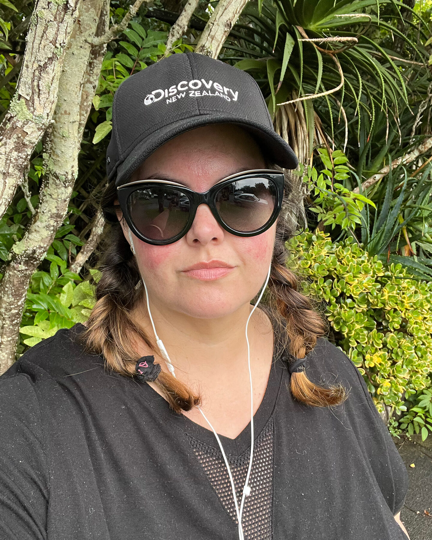 A selfie of a Māori woman walking outside in front of a garden. She is wearing a black t-shirt with sunglases and a black cap that says Discovery New Zealand. She does not look impressed.