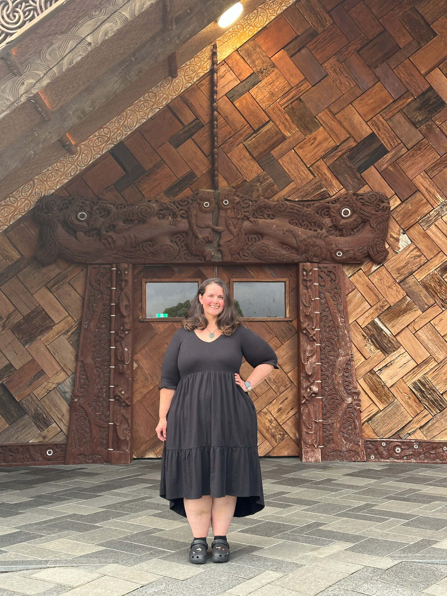 Meagan, a fat Māori woman with lipoedema, stands in front of Ngākau Māhaki, the whare whakairo (carved meeting house) on Te Noho Kotahitanga Marae. She is wearing a black flowing dress and black platform crocs.
