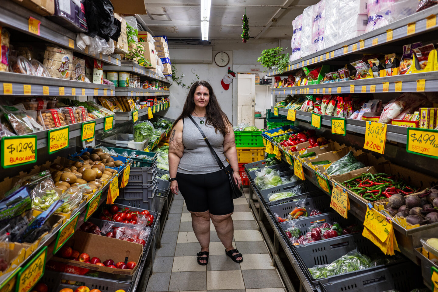 A fat Maori woman stands in the aisle of a fruit and vegetable shop.
